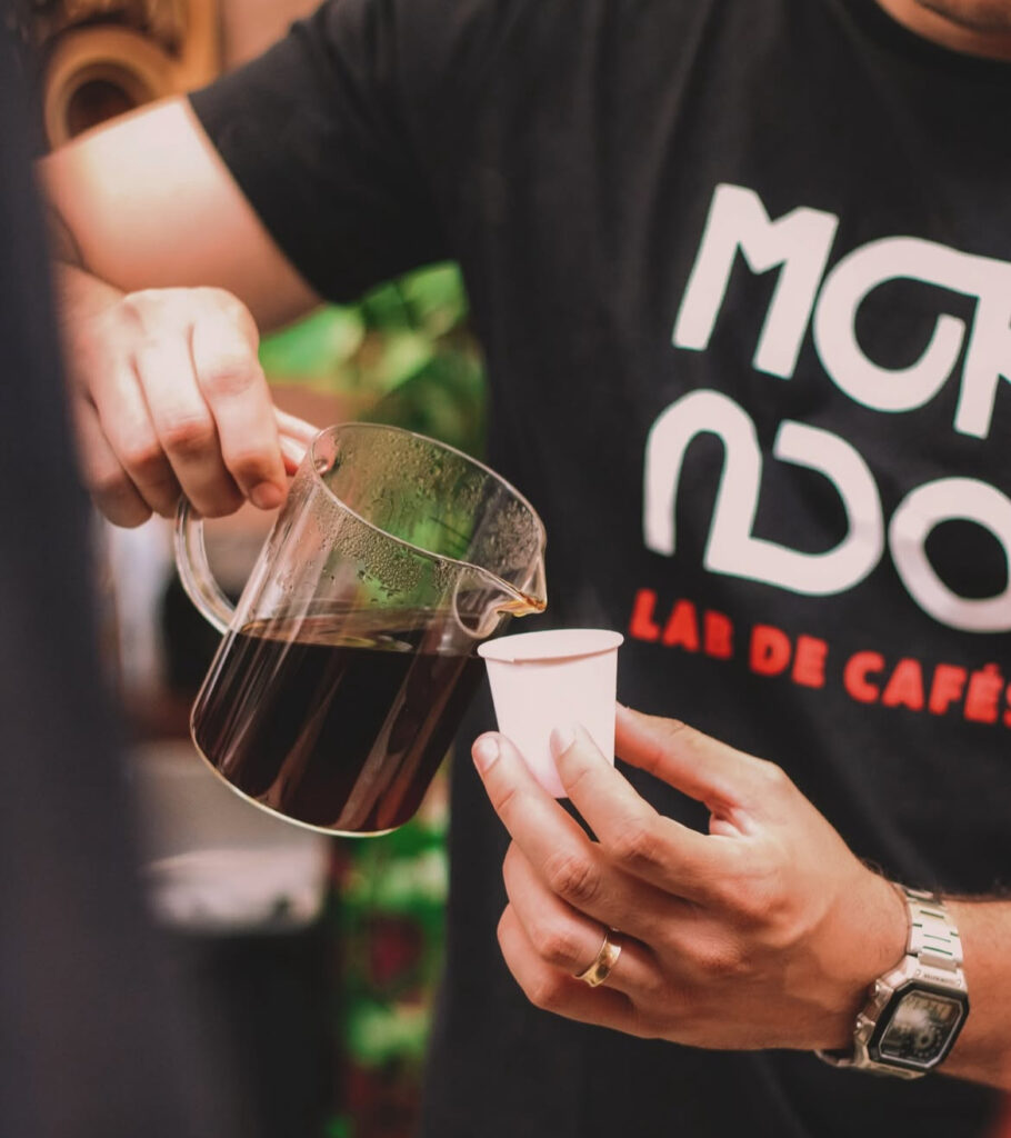 Pessoa servindo café em um copo pequeno branco de plástico com uma camiseta preta da Mokado Lab de Cafés 